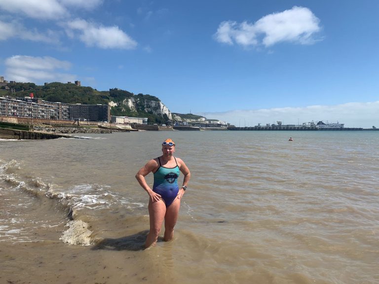 Woman in a swimsuit standing in shallow water at a beach with cliffs and a harbor in the background